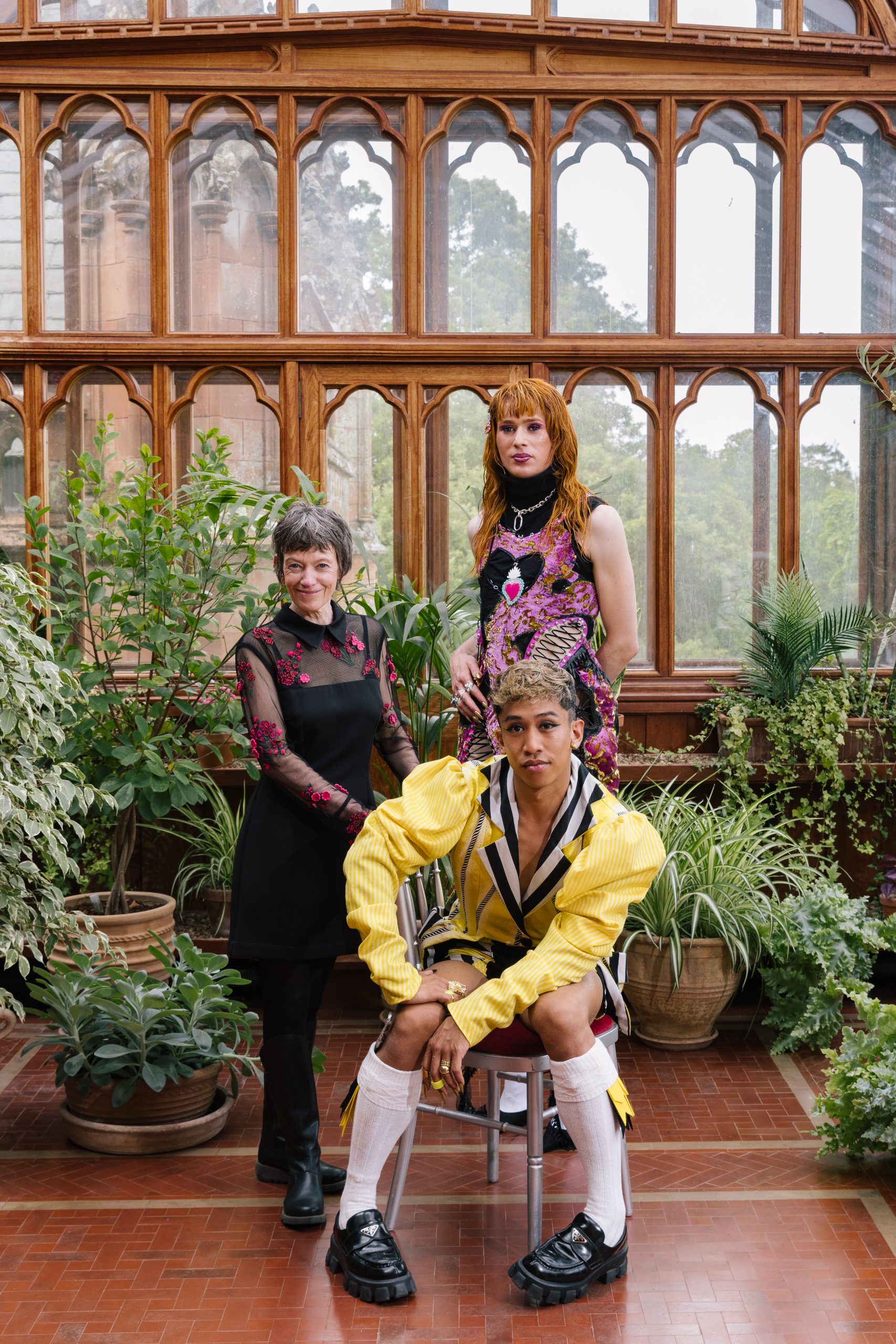 Three people pose looking towards the camera, Davide is seated in the foreground wearing puffed yellow sleeves, knee length socks and black loafers; Morven to the left wears a black dress with pink floral applique and behind Angel stands wearing black and pink with a high colour and bright heart in the center of her chest. They are in a conservatory, with wooden window frames and terracotta tiled floor, surrounded by green leafy plants.