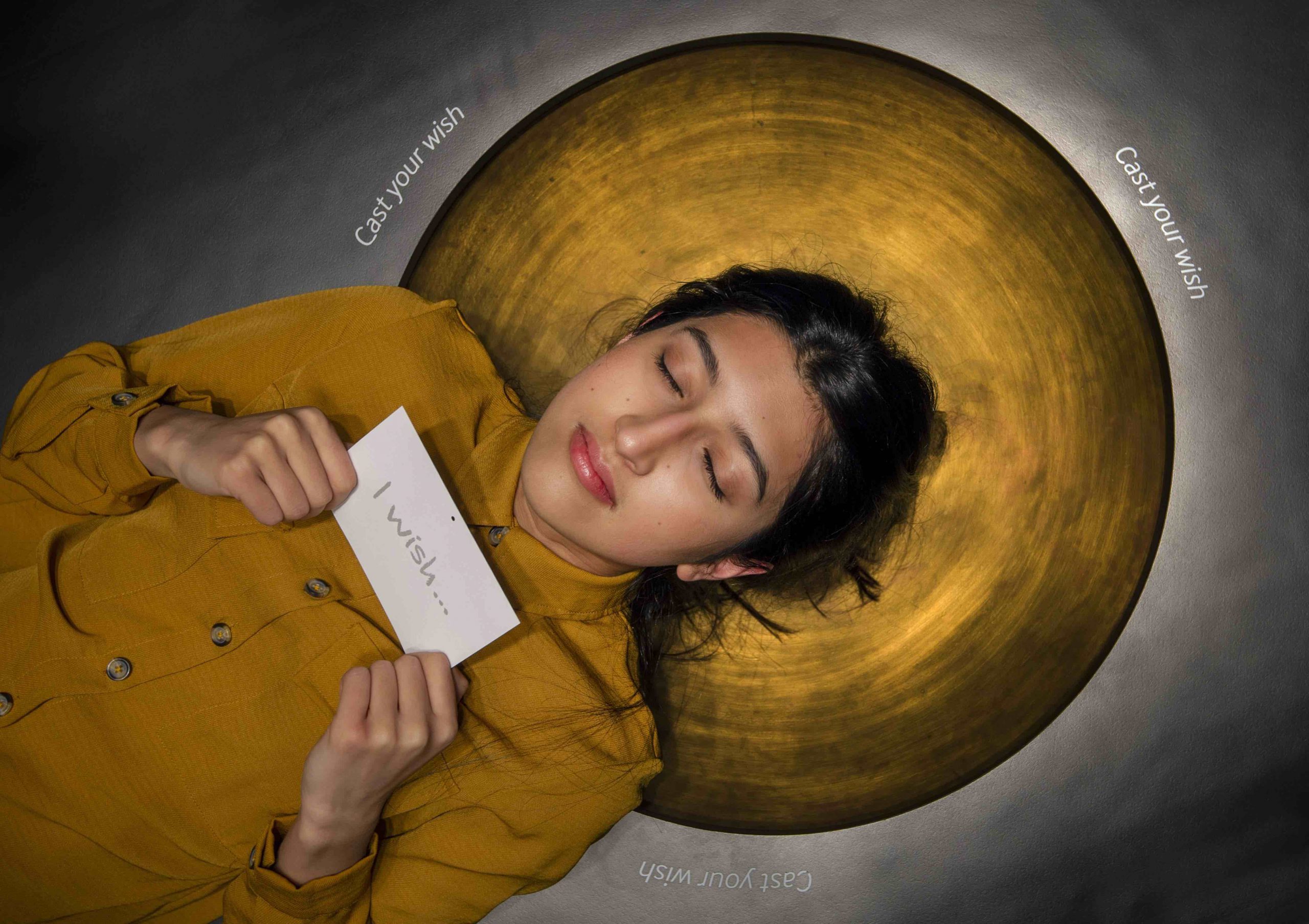 A young woman with dark hair rests on a black wishing well with a golden circle around her head. Her eyes are closed and she holds a card with the words I wish on her chest.