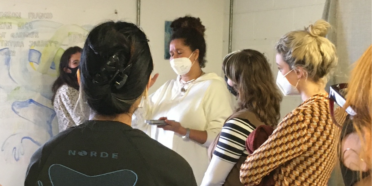 PDP participants listening to artist Alberta Whittle discuss her work at her studio in Glasgow Sculpture Studios. Alberta is showing a watercolour painting in hues of green, blue, yellow and black.