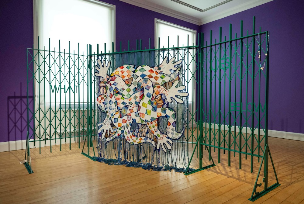 A large tapestry composed of imagery of snakes, water, diamonds and hands, suspended on a green gate